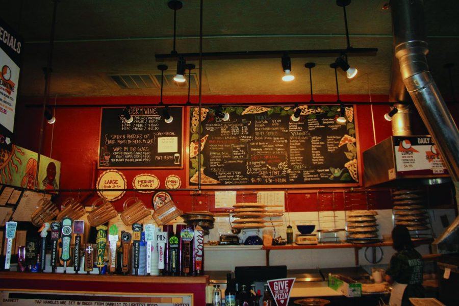 At the ordering counter in American Dream Pizza, there are drinks on tap, pizzas in various stages of production, and a large chalkboard menu. 