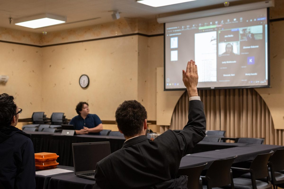 A man sits at a table with a laptop in front of them and raises their right hand in a signal to speak.