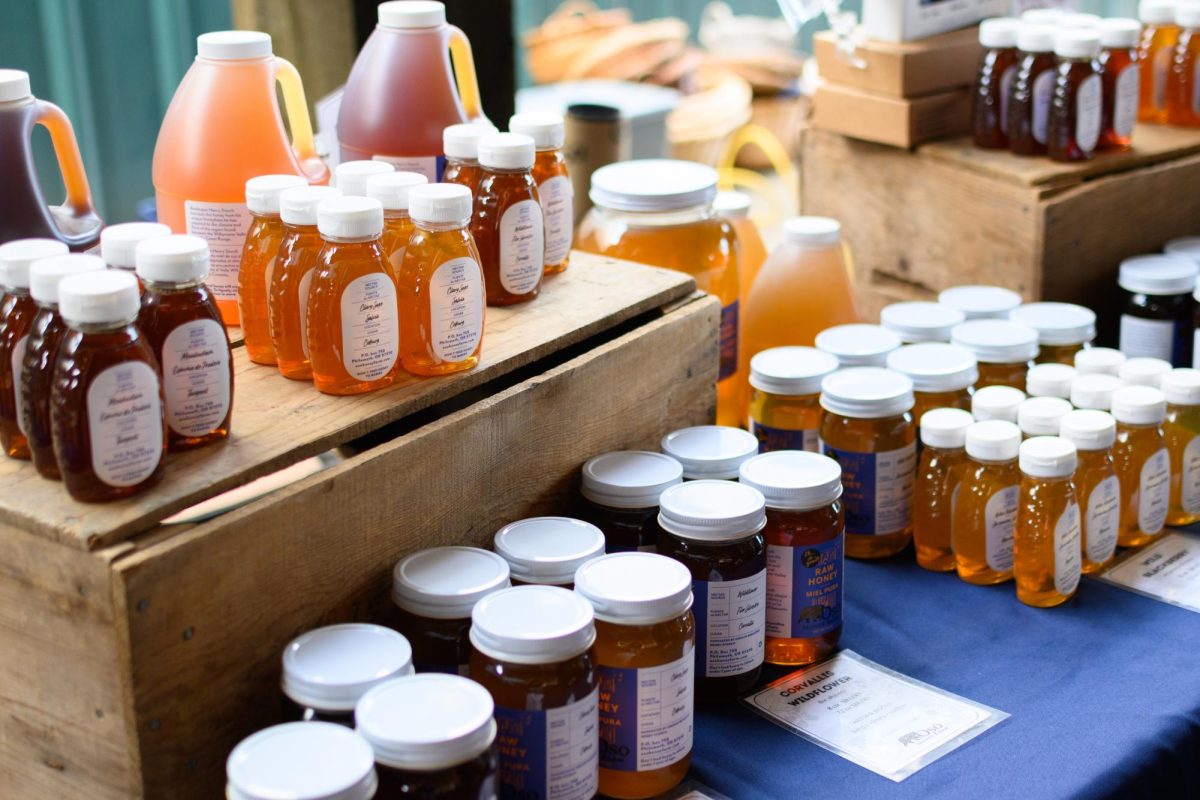 A table covered in a blue tablecloth has honey jars laid out in rows and a sign that reads "Corvallis Wildflower" from OSO Honey farm.