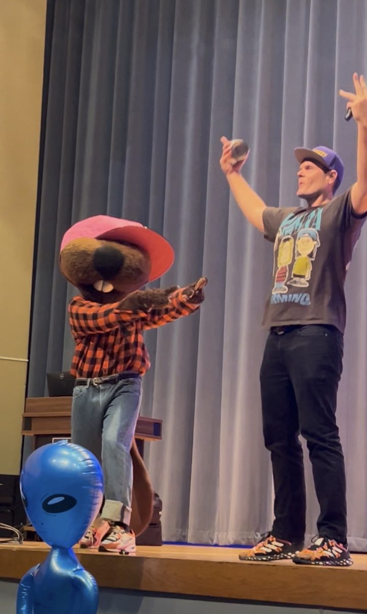 An adult man celebrating with his hands up. to his left, a person dressed as a beaver wearing a large hat points at him endearingly.