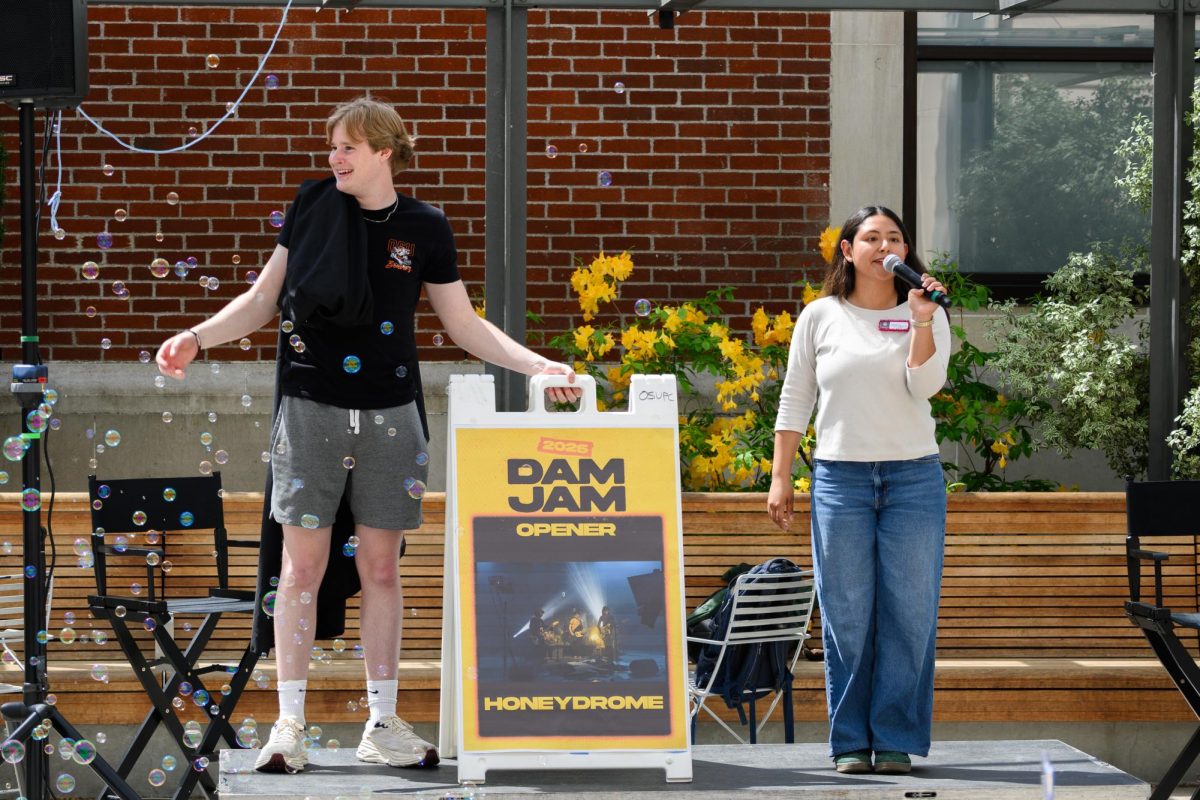 Two young adults stand on either side of the a sign saying "Dam Jam opener Honeydrome." The person on the right holds a microphone.