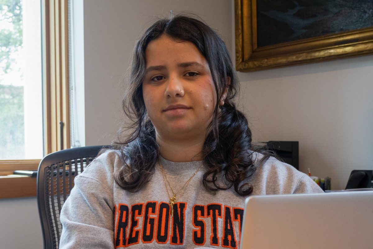 Person with black hair and grey sweater sits in an office with a laptop open.