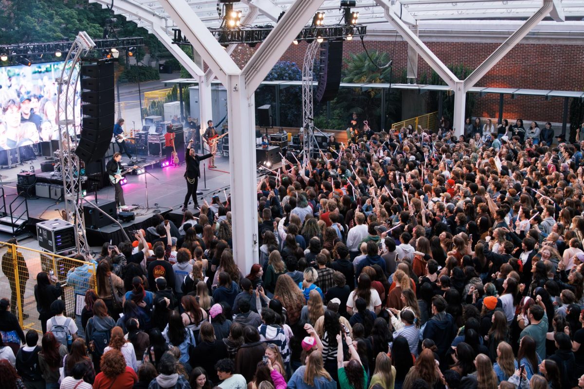 A large crowd of adults crowd an outside venue. The stage facing the audience has five men on it.
