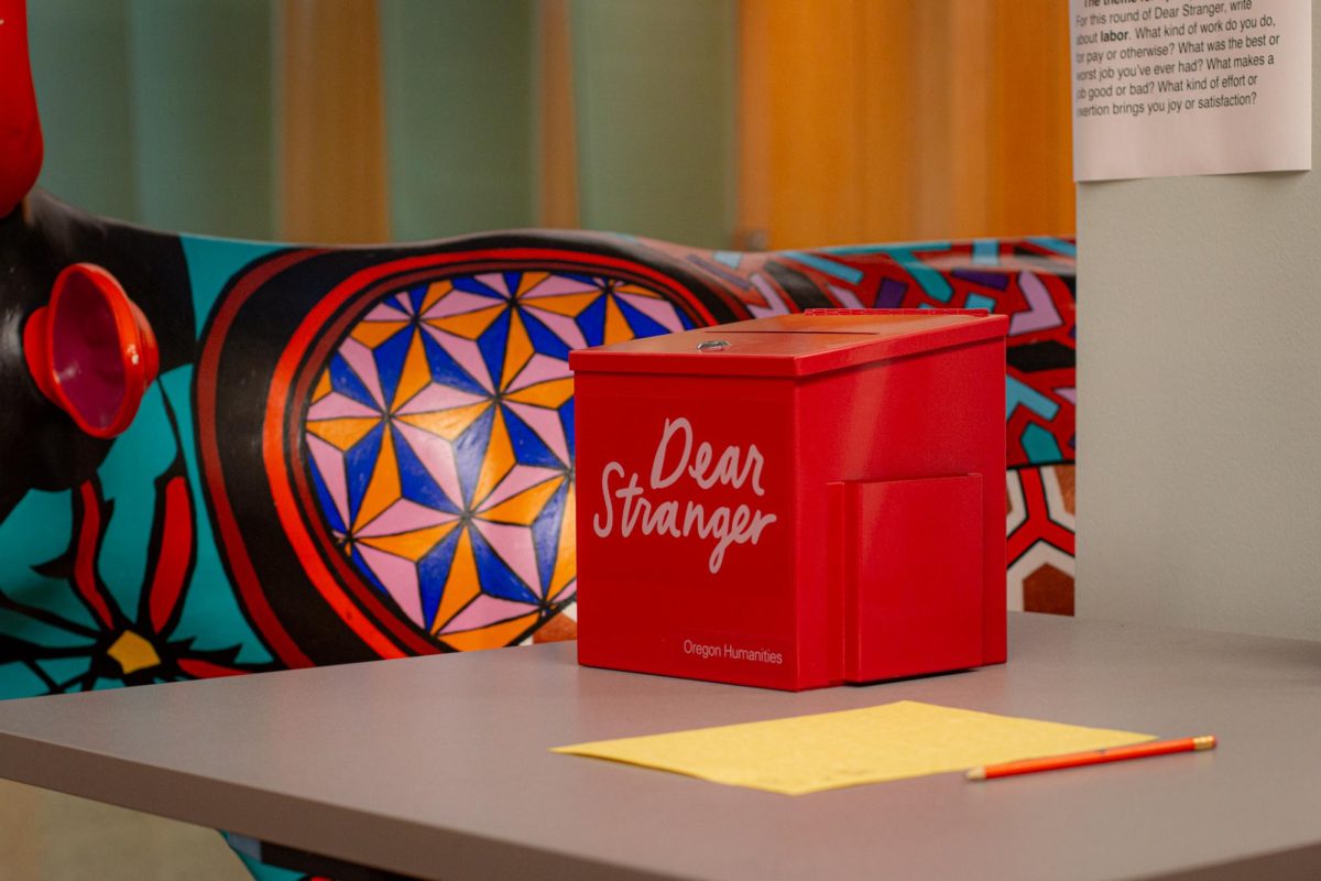 A red postal box with "dear stranger" printed the front sits on a desk, on it's right is a blank piece of paper. On it's left, is a statue of a cow covered in colorful patterns.